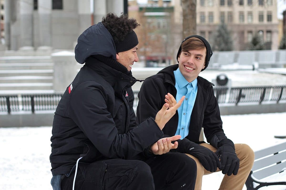 Zwei junge Männer sitzen im Schnee auf einer Bank, der eine erzählt mit den Händen, der andere hört ihm lächelnd zu