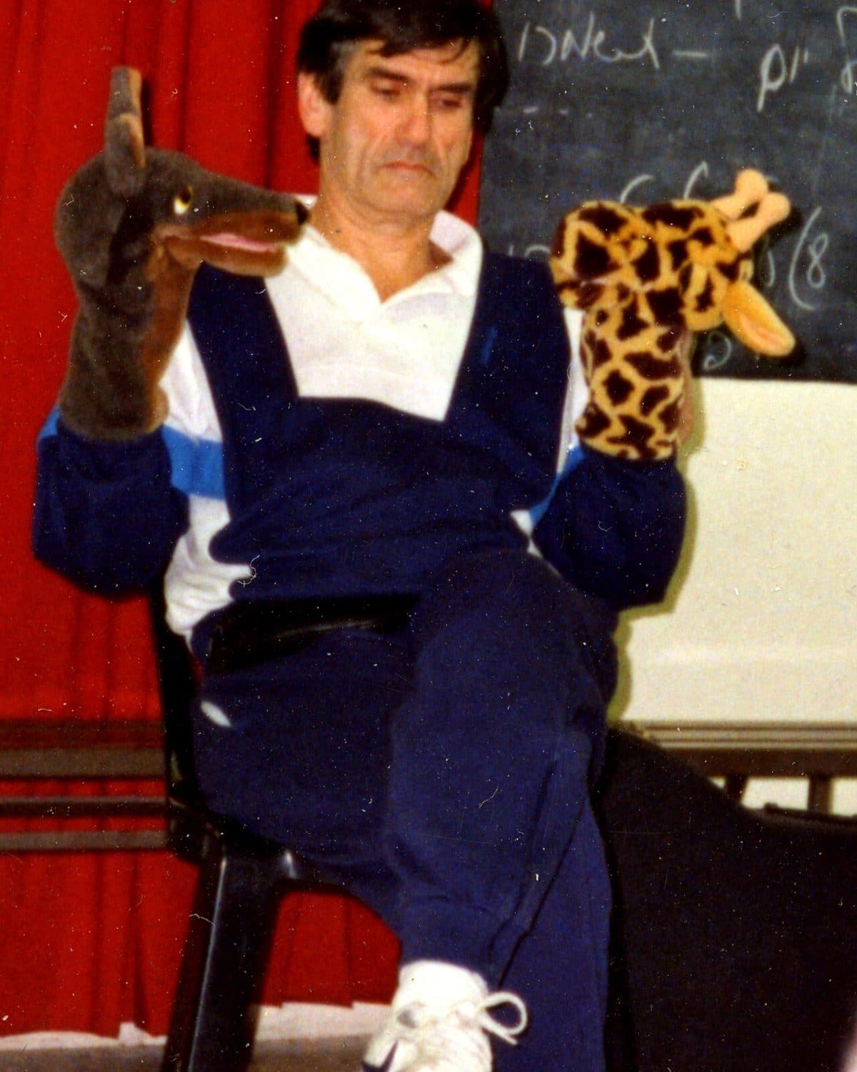 Ein Mann sitzt auf einem Stuhl vor einer Tafel und hält an der einen Hand eine Wolfspuppe, an der anderen eine Giraffenpuppe