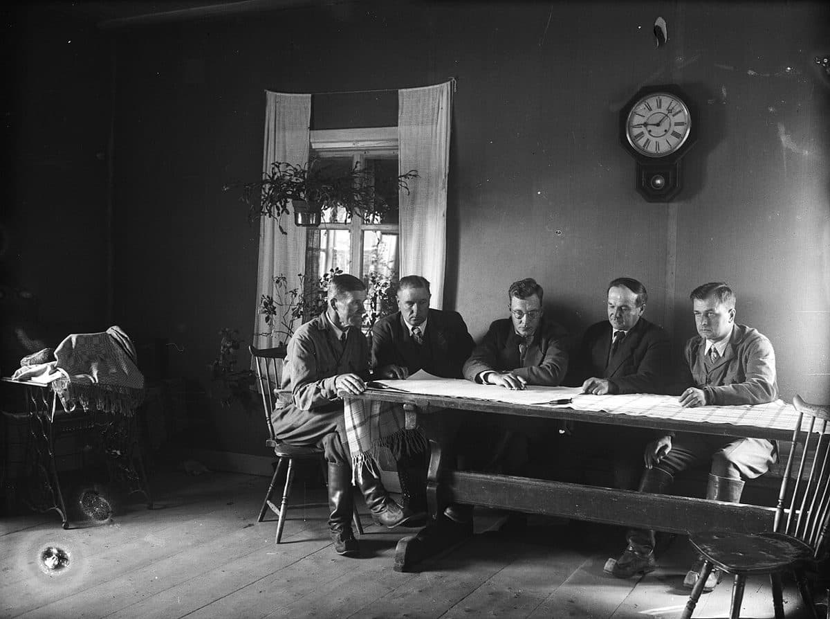 Schwarzweißfoto von fünf Männern, die an einem langen Holztisch nebeneinander sitzen und gemeinsam auf ein ausgebreitetes Papier sehen