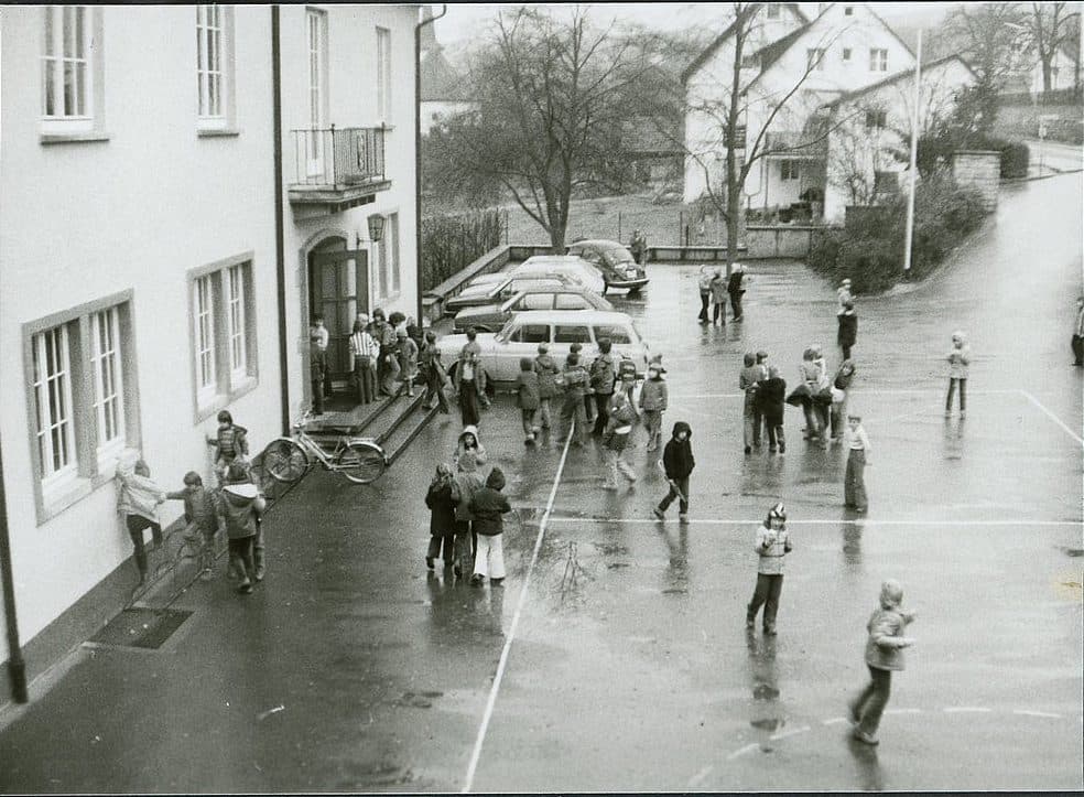 Schwarzweißfoto eines nassen Schulhofs von oben, Kinder in Winterjacken stehen in Gruppen vor dem Schulgebäude